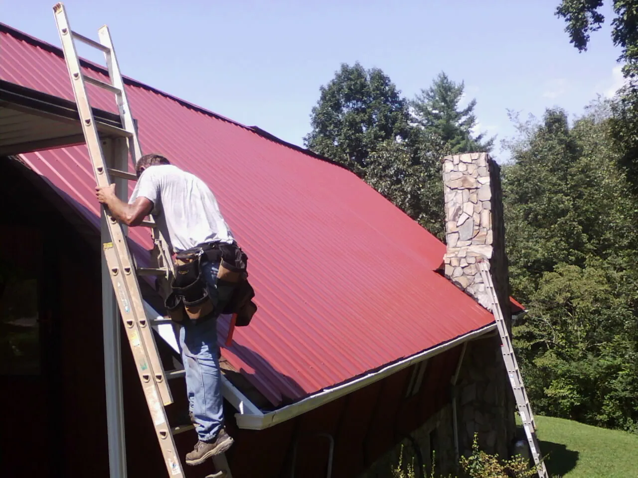 Roofer on a ladder beside a red metal roof and stone chimney — Soffit & Fascia Repair by Roofing Brothers in New Cumberland